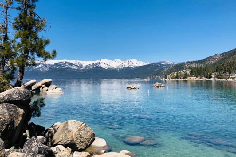 Clear blue lake with rocky shoreline, pine trees, and snow-capped mountains in the background