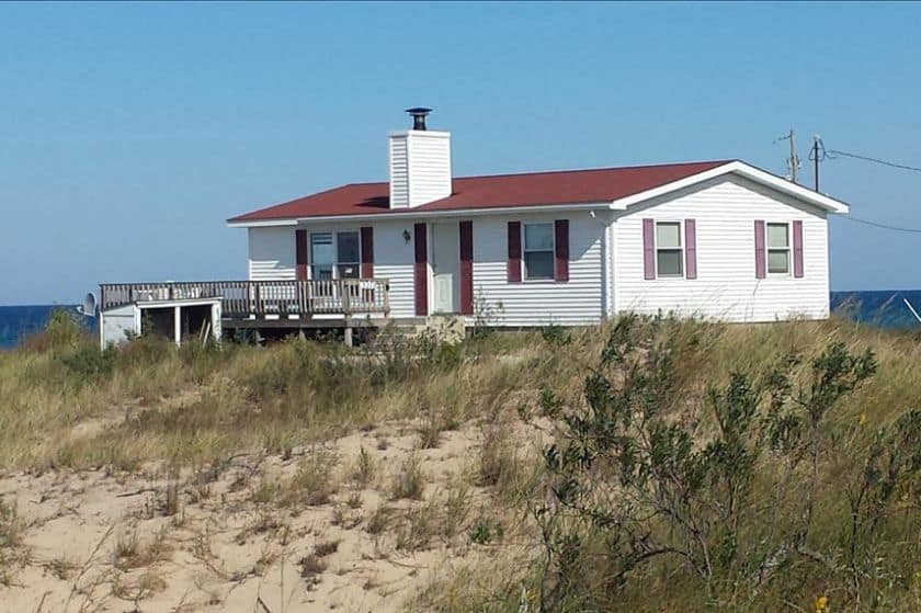 White beach house with a red roof and deck, surrounded by sand dunes and overlooking the ocean.