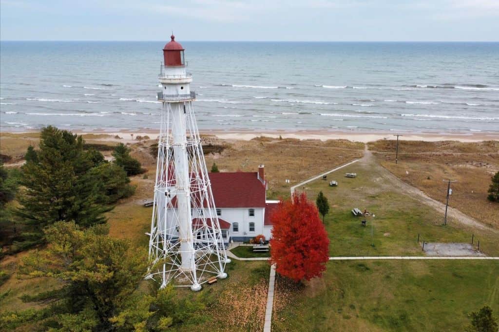 White metal lighthouse with a red roof, set near a sandy shoreline with trees and picnic tables