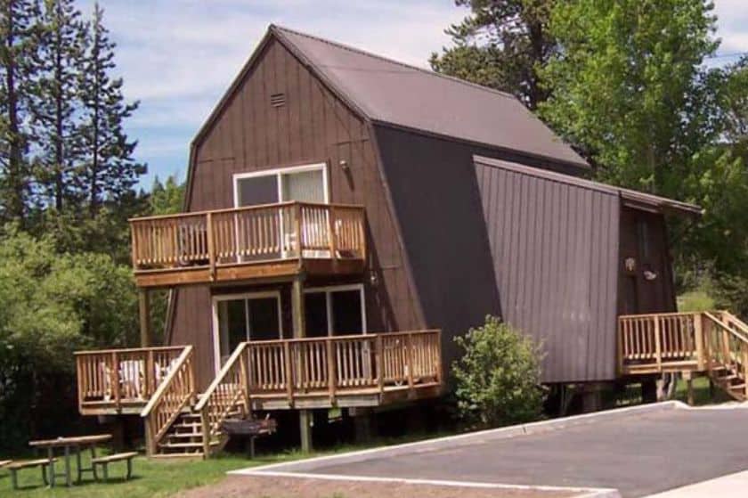 Dark brown A-frame cabin with wooden balconies and decks, surrounded by trees and a paved driveway.