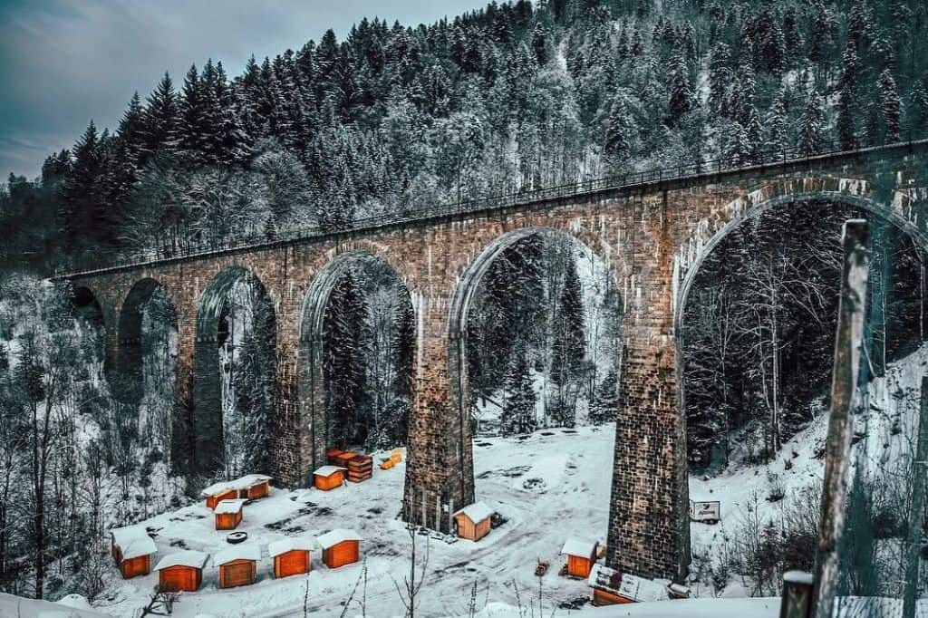  A historic stone aqueduct covered in snow, surrounded by a dense forest in winter. Small wooden cabins are scattered below the structure.