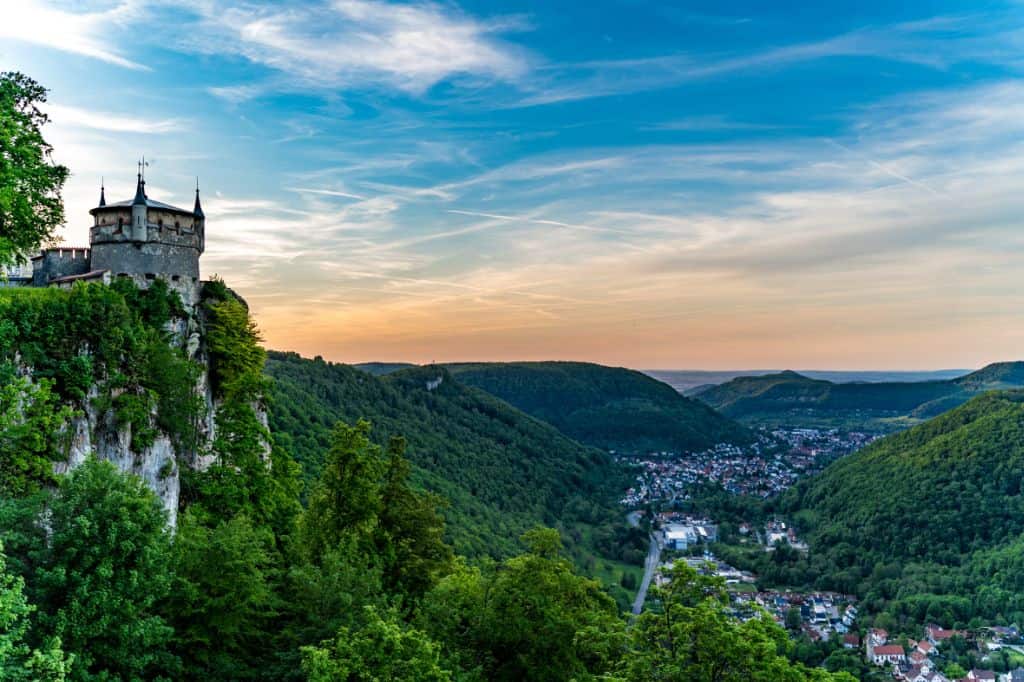 A castle perched on a cliff, overlooking a valley with a town below, under a sunset sky.