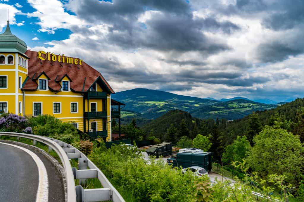  A scenic countryside view with rolling green hills, a yellow European-style building labeled "Oldtimer," and a winding road in the foreground.