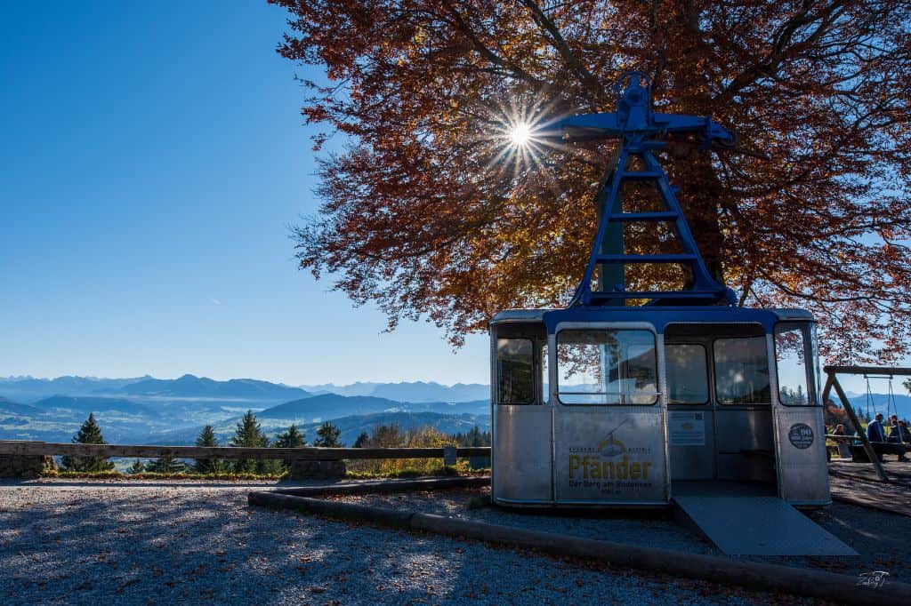 A cable car station on a mountain peak, with a bright blue sky, autumn foliage, and panoramic views of the surrounding valleys.