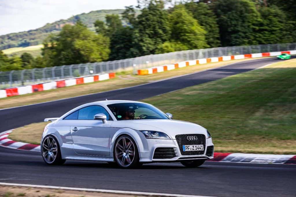 A white Audi sports car speeding around a race track with red and white curbs, surrounded by lush greenery.